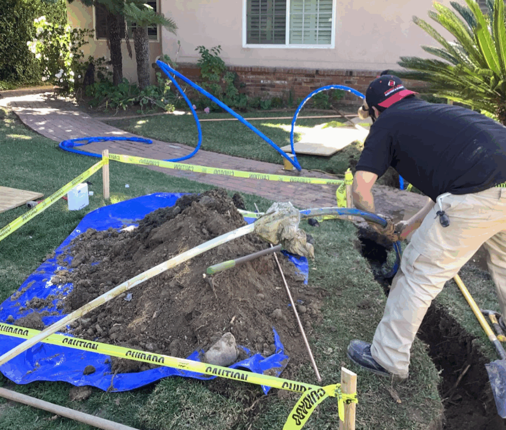 A worker in a black shirt digging a trench in a residential front yard.
