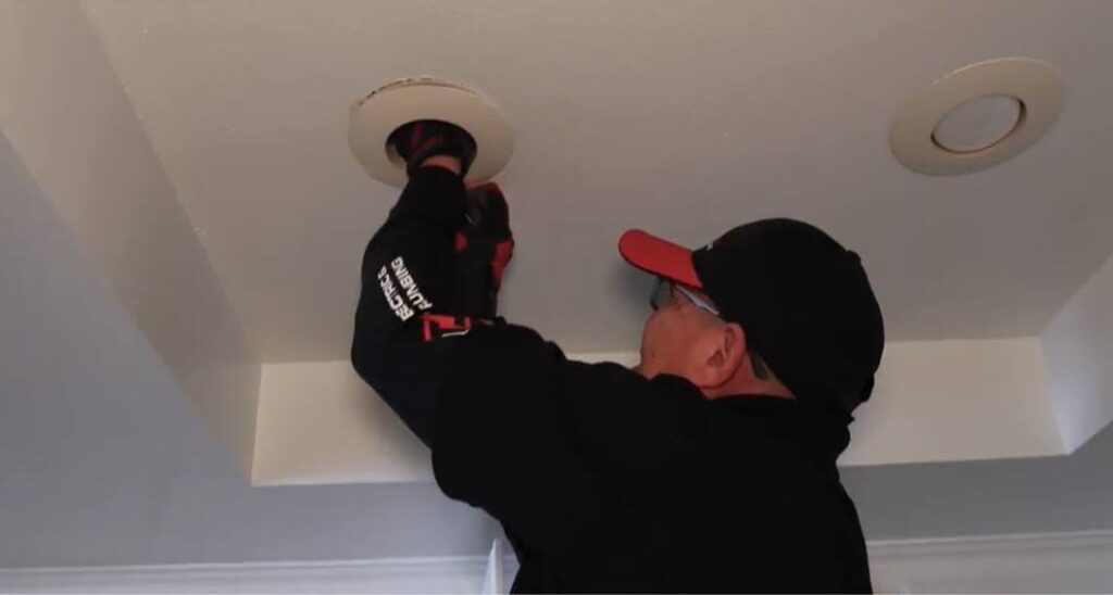 Electrician in black uniform installing ceiling light fixture.