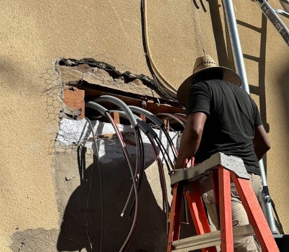 Worker on ladder installing electrical conduits through exterior wall.