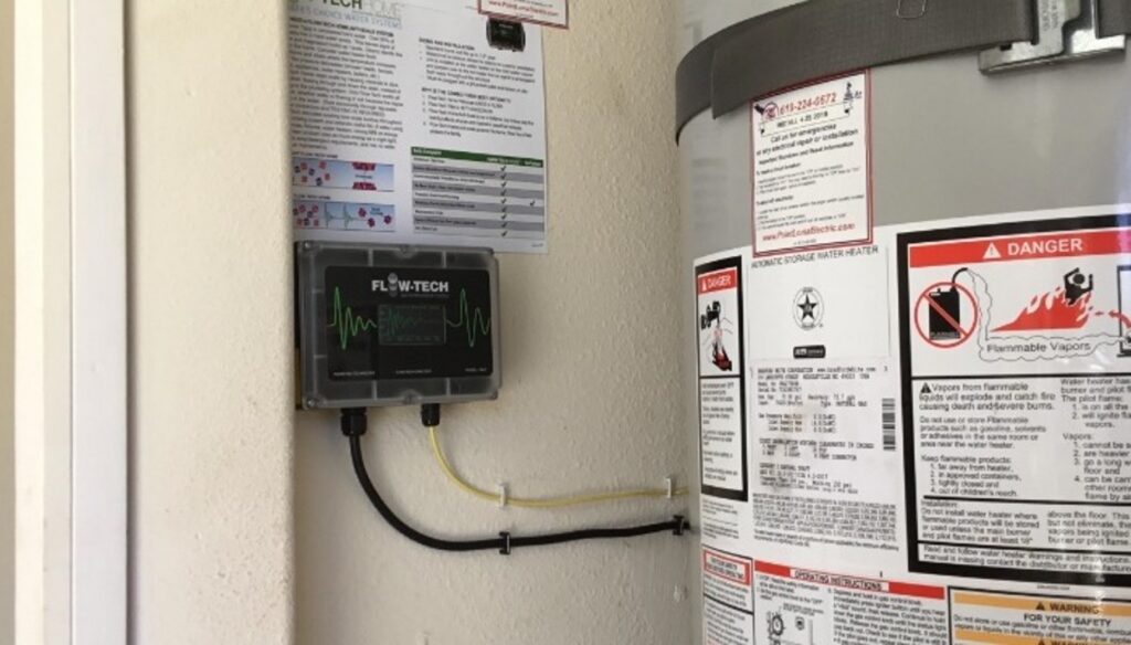 A black Flow-Tech water treatment device mounted on a wall next to a white tank water heater