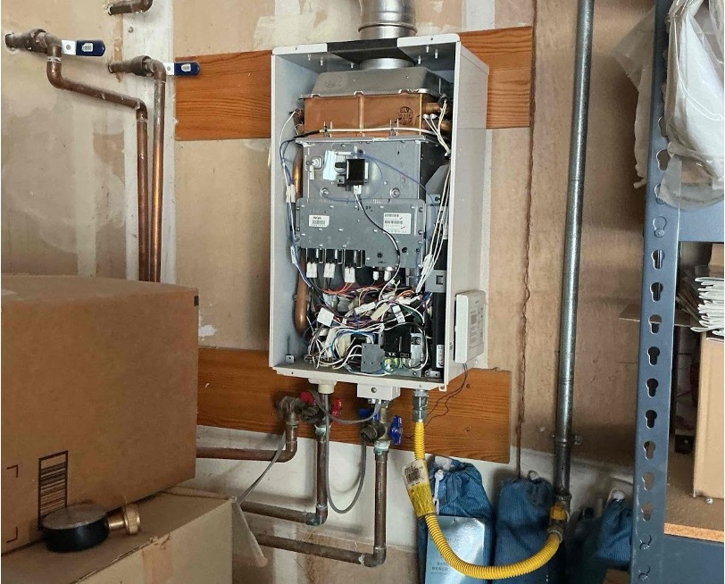 Open tankless water heater with cover removed, showing internal components, copper pipes, yellow gas line, and metal exhaust vent