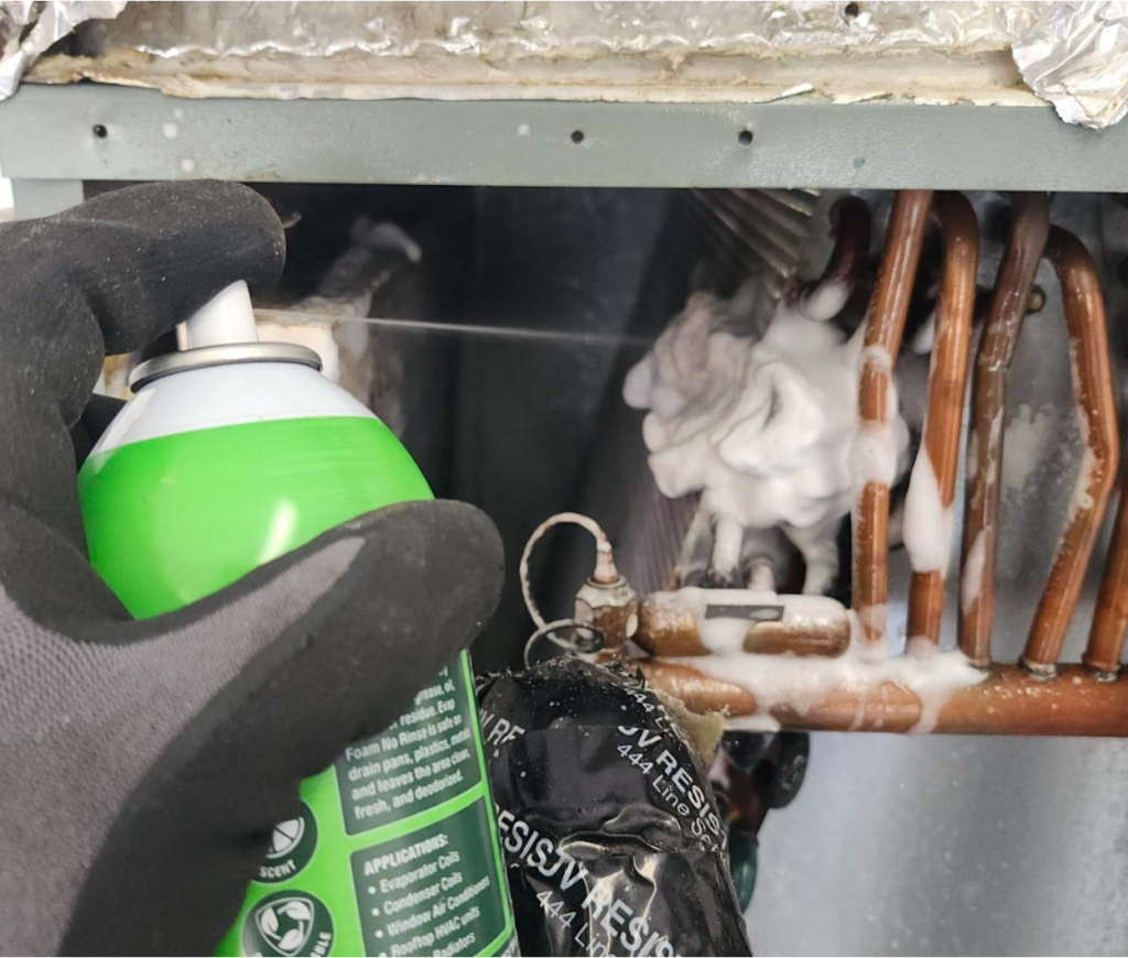 A gloved technician is spraying no-rinse foaming coil cleaner onto a copper evaporator coil tubing