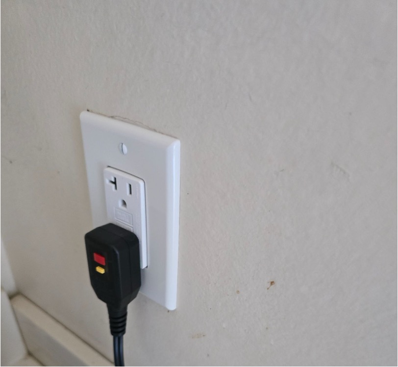 A wall outlet with a black appliance plug connected, showing red and yellow test and reset indicators