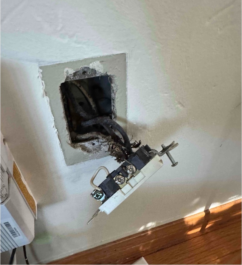 Damaged electrical outlet pulled from a wall, revealing burned, frayed wires and a blackened junction box