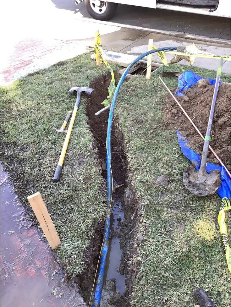 Open trench in a front yard exposing a sewer line with tools and caution tape around the excavation site