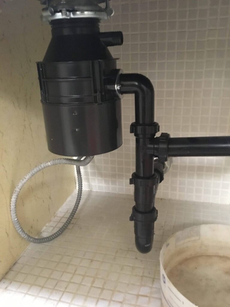 Garbage disposal mounted under a sink with a metal-clad wiring whip running into its base and the drain trap alongside.