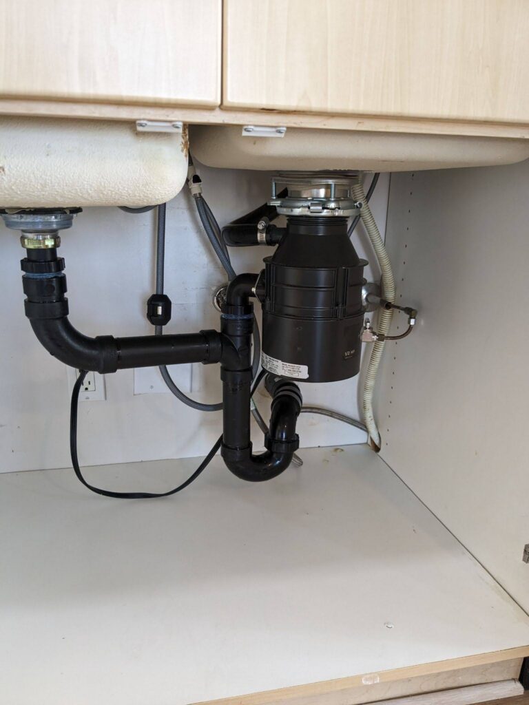 Under-sink garbage disposal and drain plumbing beside a wall outlet with appliance cords plugged in.