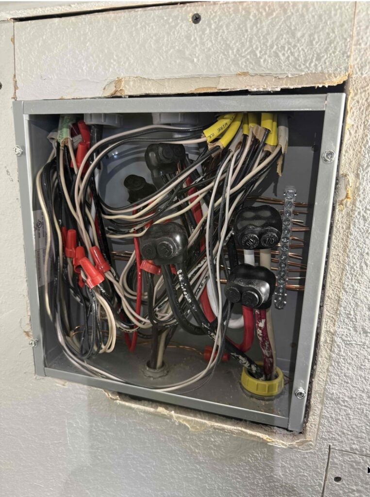 Open residential electrical panel with tangled wiring, wire nuts, and ground bus bars mounted in a wall.