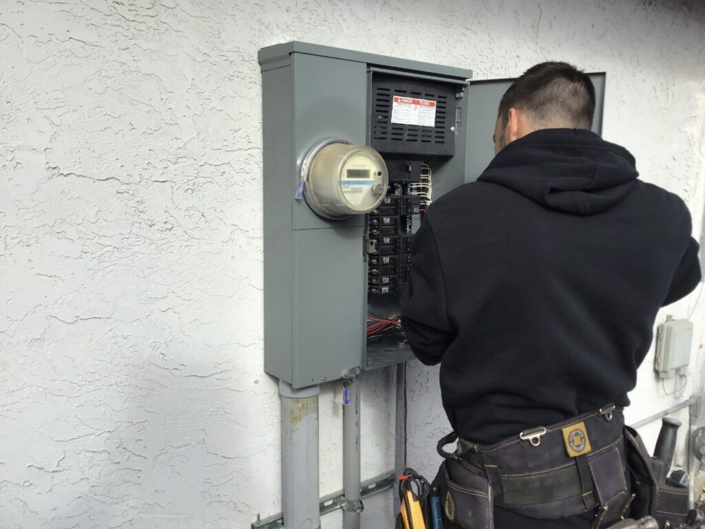 A Point Loma Home Pros electrician in a tool belt working on an open electrical panel with a digital meter.